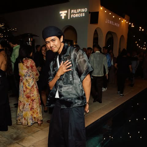 Producer, Lasi of Bolo Music wearing a black patterned short-sleeve shirt, black wide-leg pants, and a black cap stands in front of a pool at an evening event. The background shows a building with 'Filipino Forces' and 'Jeweler' signage, with other guests and warm string lights visible.